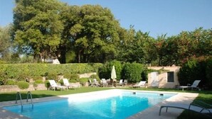 A rectangular swimming pool with a clear blue water surface, surrounded by a paved deck. There are lounge chairs and umbrellas arranged around the pool area.