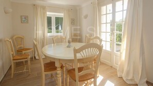 A dining room with a round table and six wooden chairs, large windows with white curtains, and a light-colored floor.