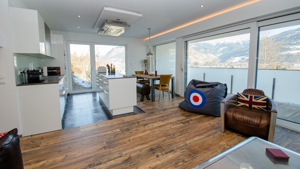 Un interior moderno con cocina, comedor y zona de estar junto a un ventanal grande con vistas a montañas.