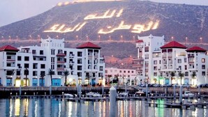 Un puerto deportivo con edificios en primer plano y una colina con escritura árabe al fondo.