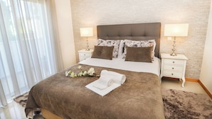 Uma cama bem arrumada com cabeceira marrom, duas mesas de cabeceira com abajures e uma janela com cortinas translúcidas.