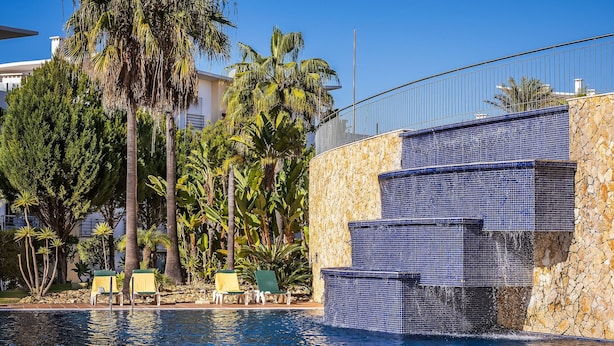 Piscina com parede de pedra e azulejos de mosaico azul, rodeada por palmeiras e espreguiçadeiras.