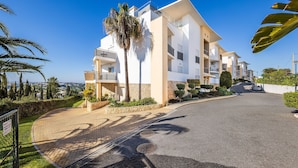 Um edifício residencial moderno, com várias andares, varandas, entrada pavimentada e uma palmeira.