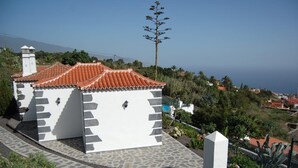 Un pequeño edificio blanco con techo de tejas, rodeado de vegetación y una vista despejada del mar.