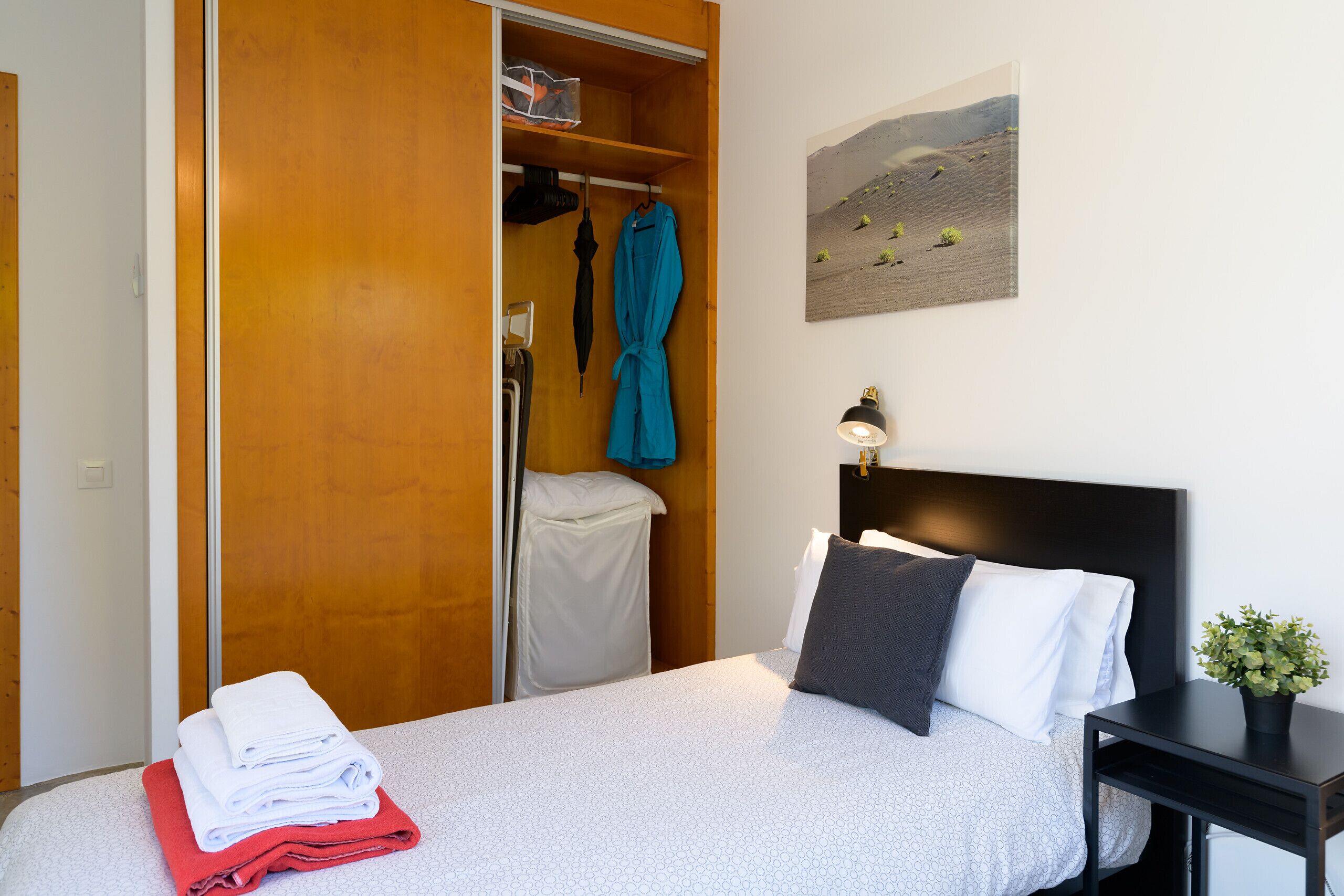 Ein Hotelzimmer mit Bett, Nachttisch mit Lampe, einem Bild an der Wand und einem Schrank mit Bademantel und Handtüchern.