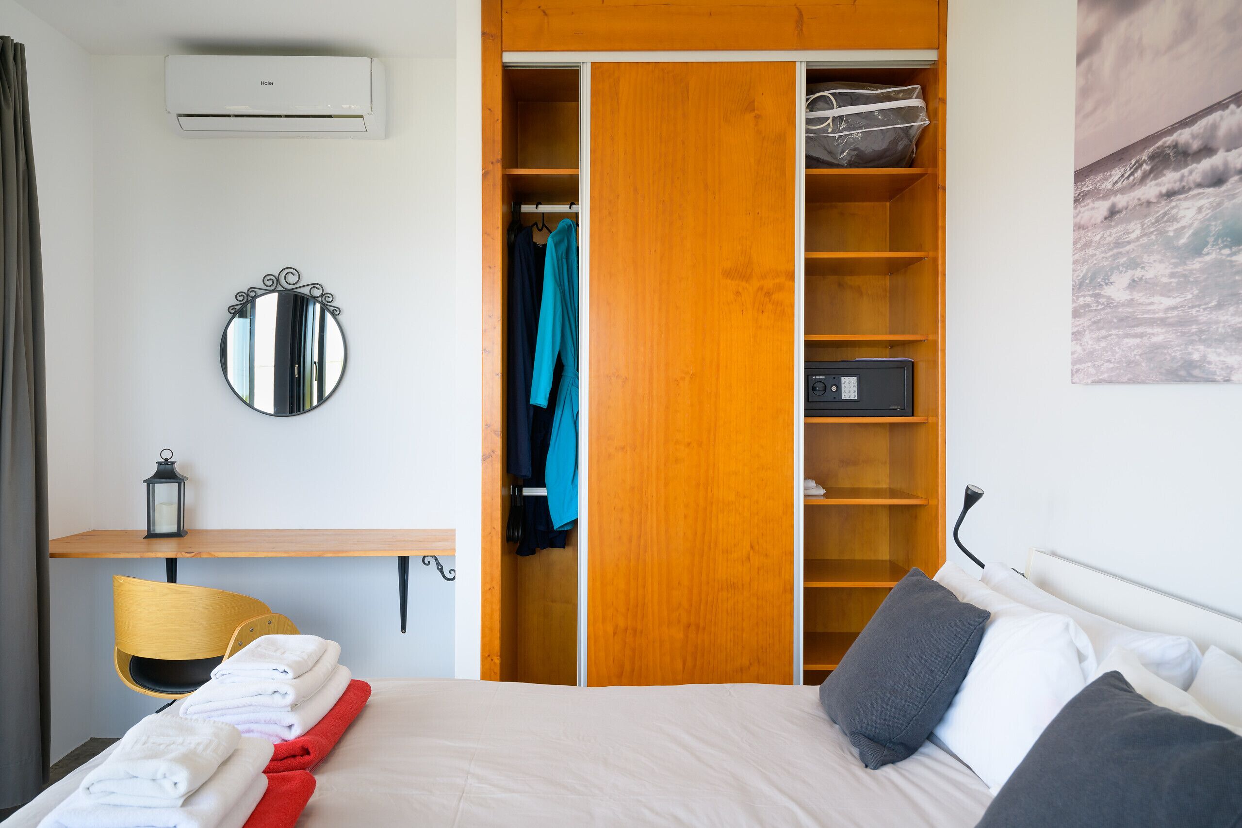A hotel room with a bed, a desk with a chair, a wardrobe, a mirror, and a wall-mounted air conditioner.