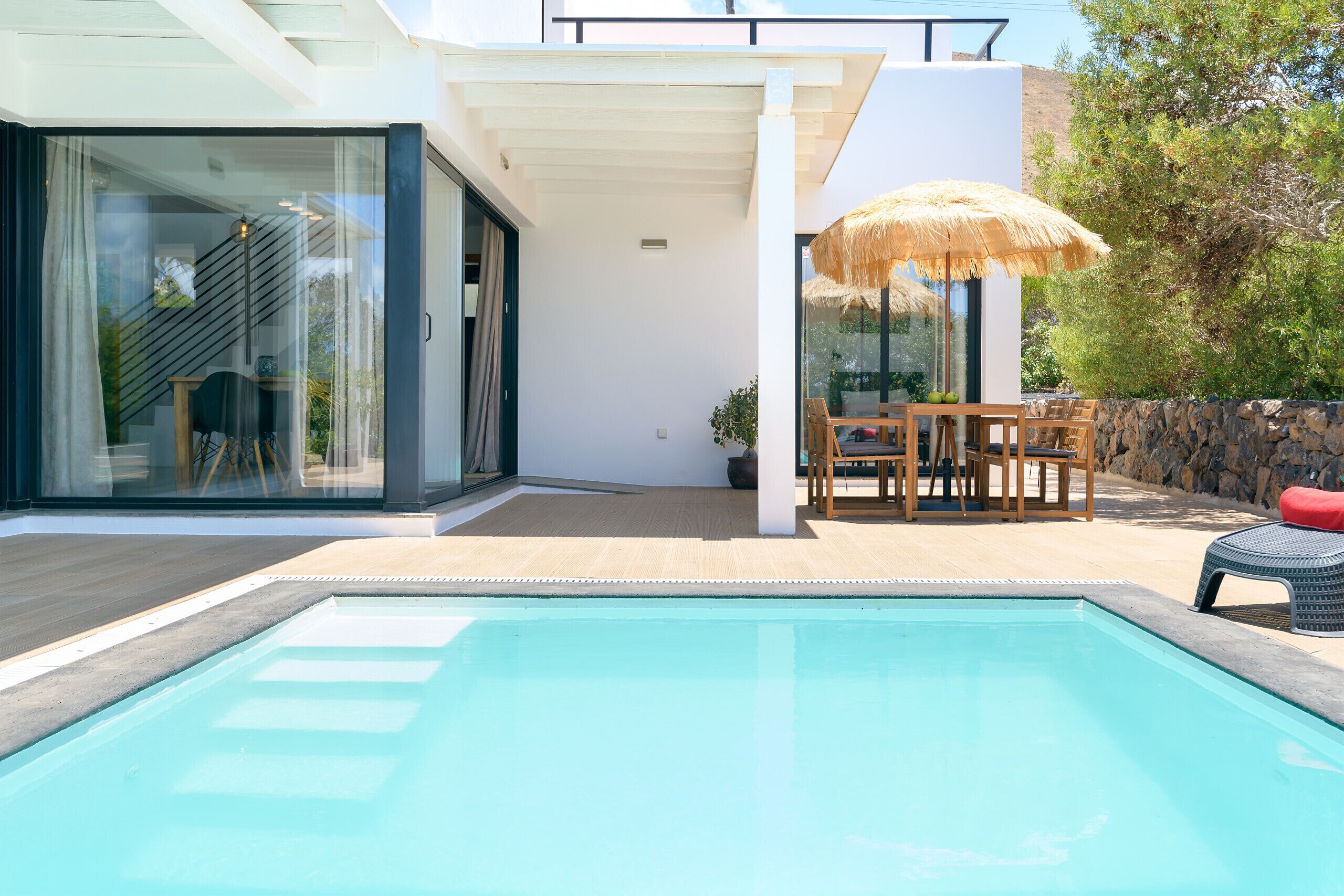 Uma casa moderna com piscina, área de refeições com mesa e cadeiras, e um grande guarda-sol.