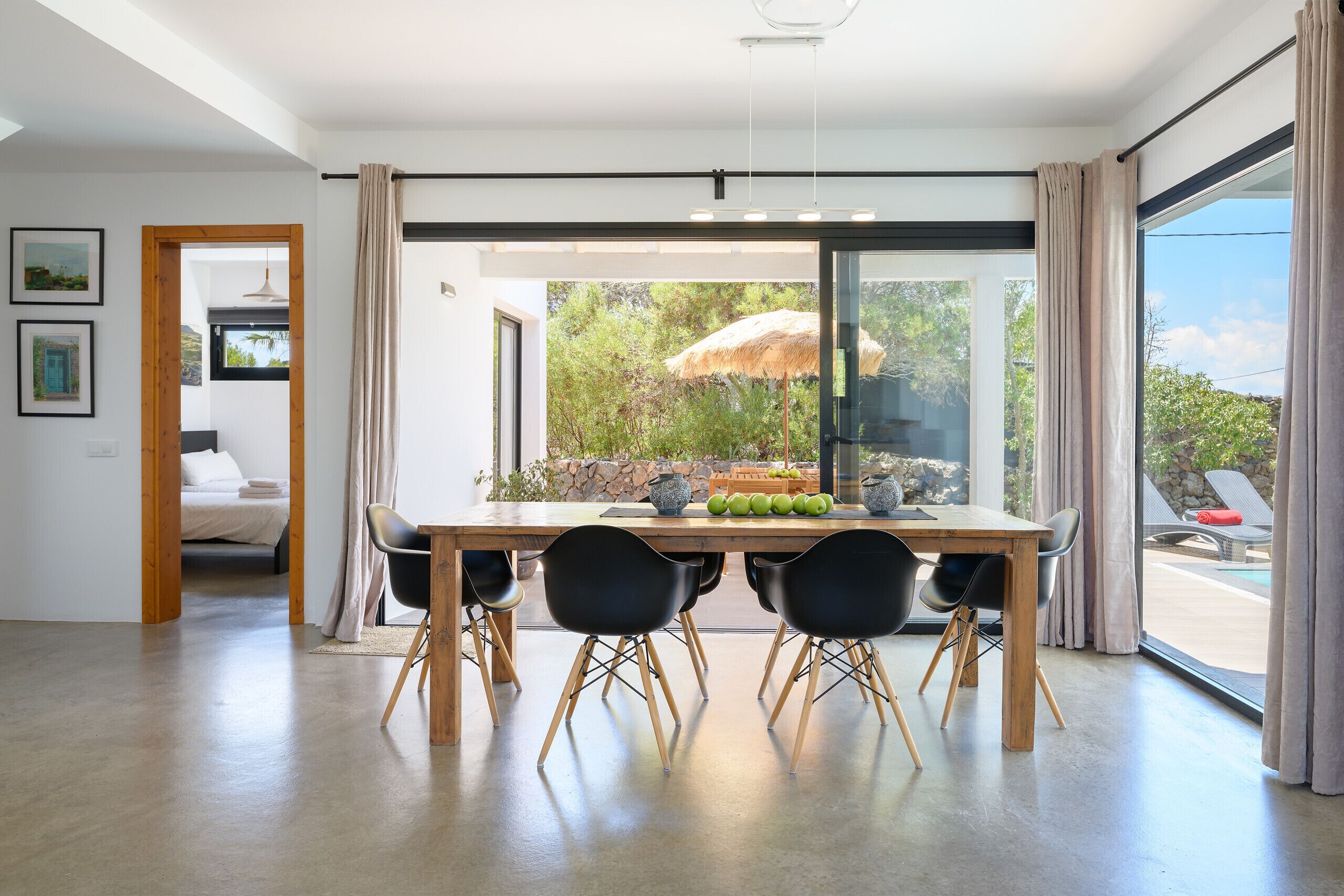 Un espace repas moderne avec une table en bois et des chaises noires, de grandes fenêtres avec des rideaux et une vue sur une piscine extérieure.