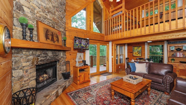 A spacious living room with a stone fireplace, wooden flooring, a large rug, leather furniture, and a balcony with a wooden railing. - Very Private with Incredible Mountain Views! Free Pool and Putt Putt Access! (Sevierville)