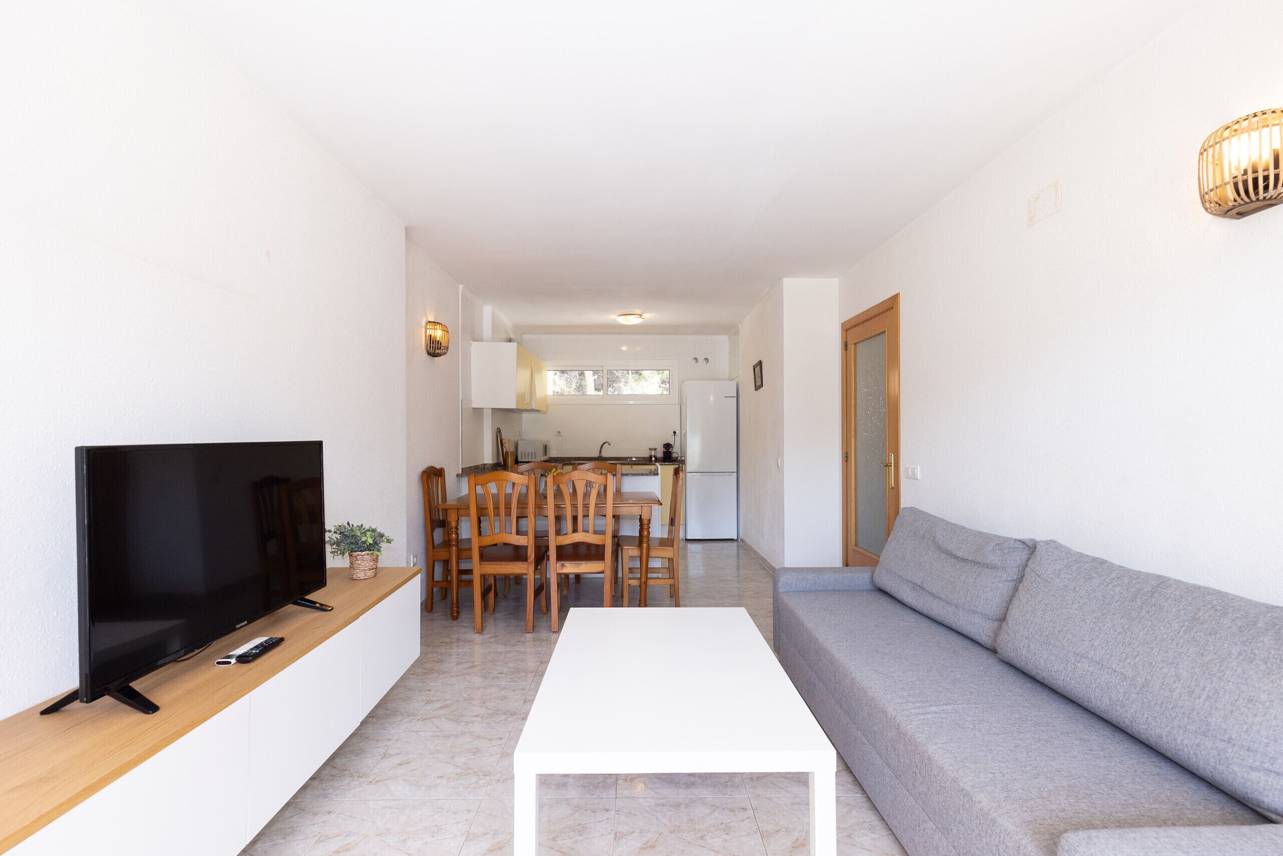 Una sala de estar con un televisor de pantalla plana, una mesa de centro blanca, un sofá gris, sillas de comedor de madera y una cocina al fondo.