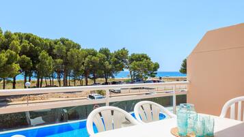 Mesa con sillas y vasos azules junto a la piscina, con vista a una playa que muestra coches y árboles.