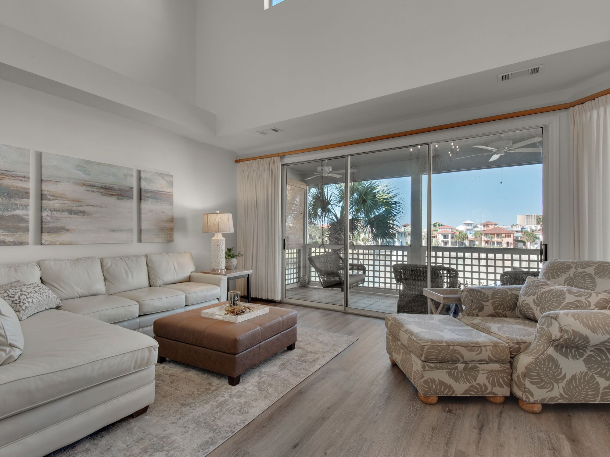 Ein geräumiges Wohnzimmer mit einem großen Schiebeglasfenster, das auf einen Balkon führt, ein beiges Sofa, ein gemustertes Pouf und ein Beistelltisch mit einer Lampe.