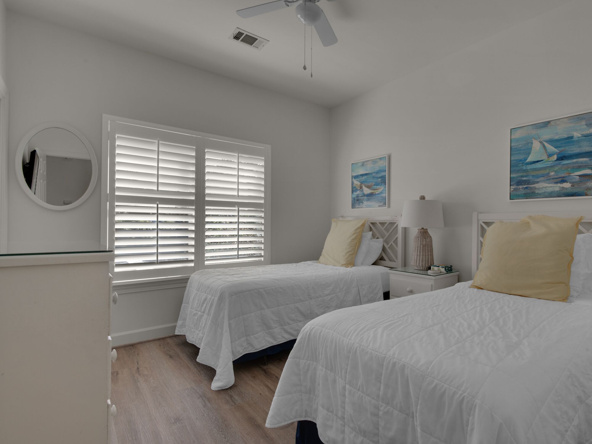 A bedroom with two beds, a ceiling fan, a window with blinds, a bedside table with a lamp, and artwork on the wall.