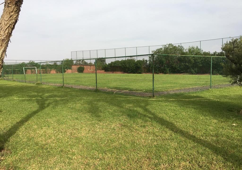 A fenced sports field with a goalpost, surrounded by trees and a clear sky.