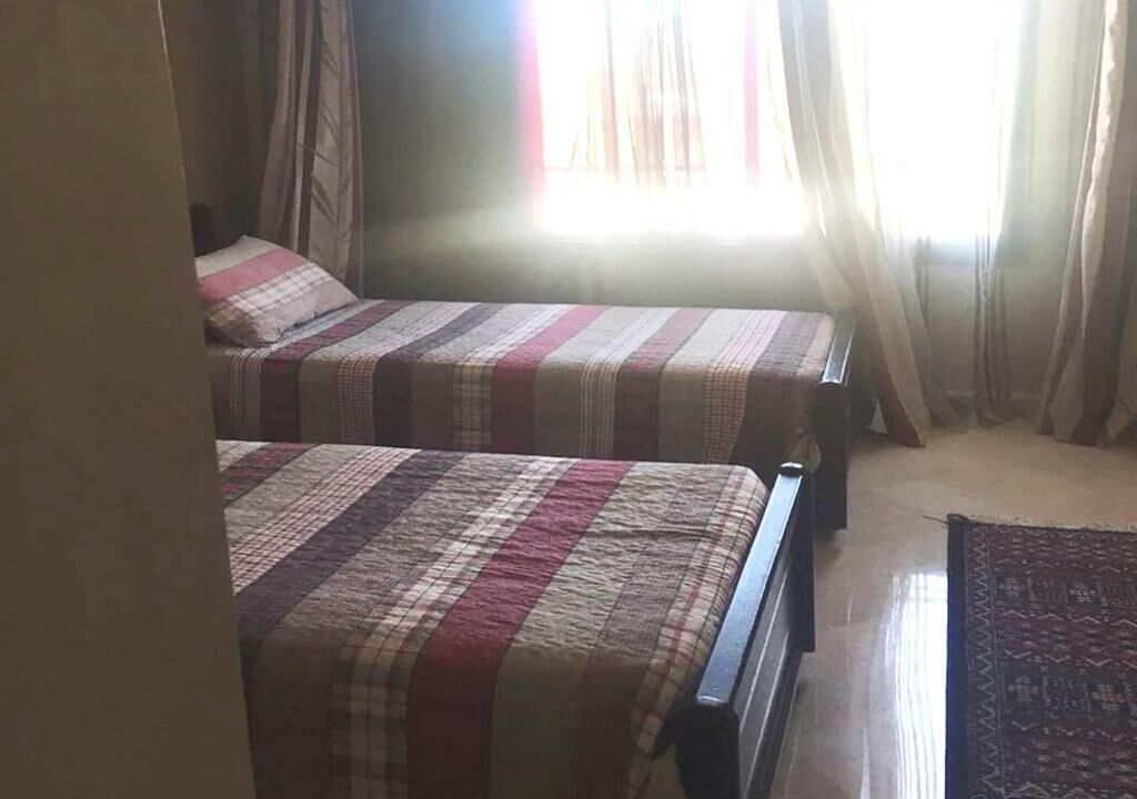 A room with two beds, each with a striped bedspread, a window with sheer curtains, and a patterned rug on the floor.
