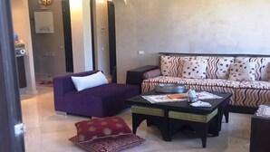 A living room with a purple sofa, a patterned sofa, a coffee table, and a red cushion. - Beautiful apartment in River Palm 4397 (Marrakech)