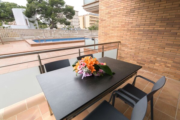 A balcony with a table and chairs, a flower arrangement, and a view of a swimming pool and buildings in the background.