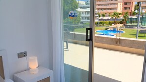A balcony with a glass door, a white bedside table with a lamp, and a view of a swimming pool and buildings in the background.