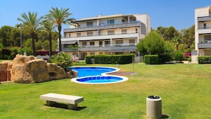 A pool area with a bench and trash can, surrounded by well-maintained grass and rocks, with a multi-story building in the background.