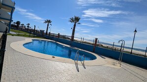 Piscina com deck revestido, cercada por palmeiras e uma grade.