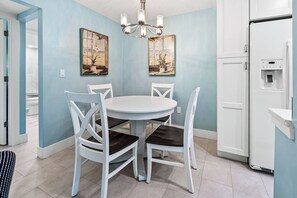 A dining area with a round table, white chairs, two wall-mounted paintings, a chandelier, and a white refrigerator.