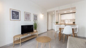 A modern living room with a TV, a round wooden table, and a dining area with white chairs. - AirCon Apartment with Pool · Beach 100m · Parking · RUISEÑORES (Salou)