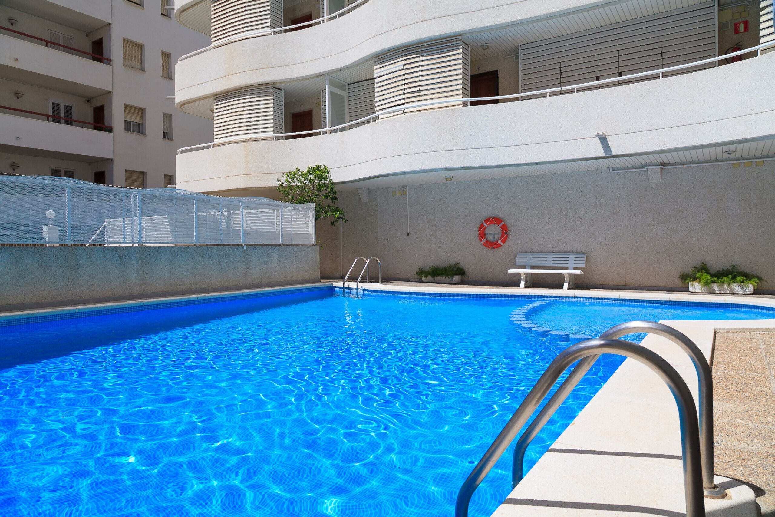 A swimming pool with a metal ladder, surrounded by a building with balconies and a lifebuoy on the wall.