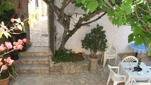 Une terrasse dallée de pierres, avec des chaises et une table en plastique blanc, entourée de plantes en pot et d’un arbre.
