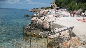 Une plage à l’eau claire, bordée de formations rocheuses et dotée d’un escalier métallique qui descend jusqu’à l’eau.