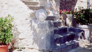 Un escalier en pierre mène à un mur de pierre surmonté d’une petite arche. Des plantes en pot se trouvent au pied de l’escalier et un poteau métallique se dresse sur la droite.