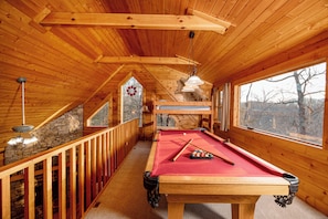 A wooden interior with a pool table, wooden railings, and large windows.