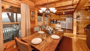 Interior de una cabaña de madera con una mesa de comedor para cuatro, una zona de cocina y un gran ventanal con vistas a un paisaje natural.