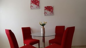 A dining area with a round glass table and four red chairs. There are two floral paintings on the wall above the table.