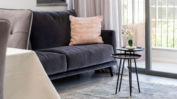 Una sala de estar moderna con un sofá gris oscuro, una almohada rosa suave y una pequeña mesa redonda con un jarrón de flores.