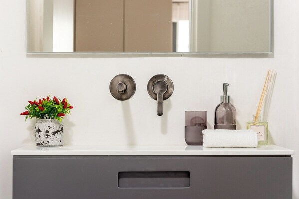 A bathroom vanity with a mirror, two faucets, a potted plant, a soap dispenser, a towel, and a bottle of reed diffuser.