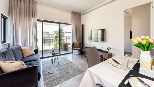 A modern hotel room with a sofa, dining table, and a balcony with a view.