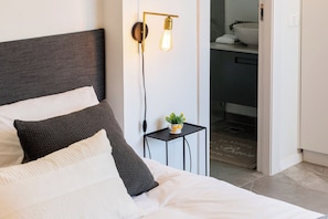 A neatly made bed with a dark headboard and white bedding, a bedside table with a small plant, and a wall-mounted light fixture.