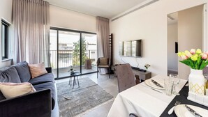 Una sala de estar moderna con sofá, una mesa de comedor para dos personas, televisor y un balcón con cortinas.