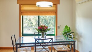 En matplats med ett glasbord, stolar, en vas med blommor, en hängande lampa och ett stort fönster med utsikt över grönområden.