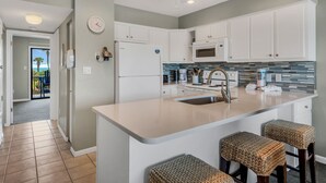 A modern kitchen with white cabinets, a stainless steel sink, and a large island with bar stools. - Two Balconies, Walk to the Beach, Pool, Beach Service, Remodeled Bathrooms (Miramar Beach)