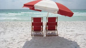 Two red beach chairs under a red and white striped beach umbrella on a sandy beach with the ocean in the background. - Two Balconies, Walk to the Beach, Pool, Beach Service, Remodeled Bathrooms (Miramar Beach)