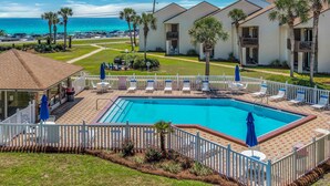 A pool area with lounge chairs and umbrellas, surrounded by a white fence, with a view of the ocean and a parking lot in the background. - Two Balconies, Walk to the Beach, Pool, Beach Service, Remodeled Bathrooms (Miramar Beach)