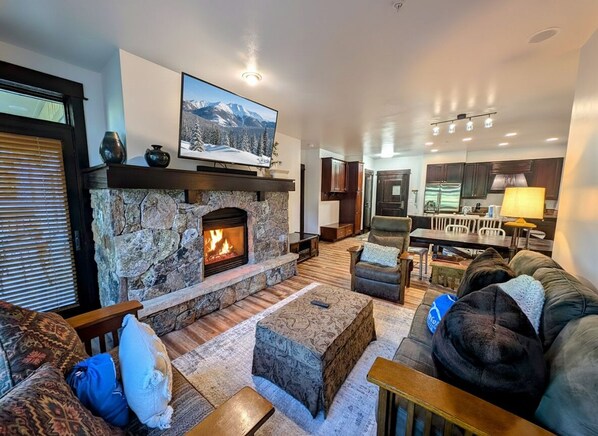 A cozy living room with a stone fireplace, comfortable seating, a television, and a kitchen area in the background. - Unit With/Private Balcony, Walk to Dining & Shops. (Keystone)