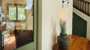Een kamer met een groot raam, een houten tafel, een kamerplant, een wandlamp en een trap.