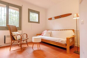 A room with a wooden sofa, a wicker chair, a small white table, a floor lamp, and a surfboard on the wall.