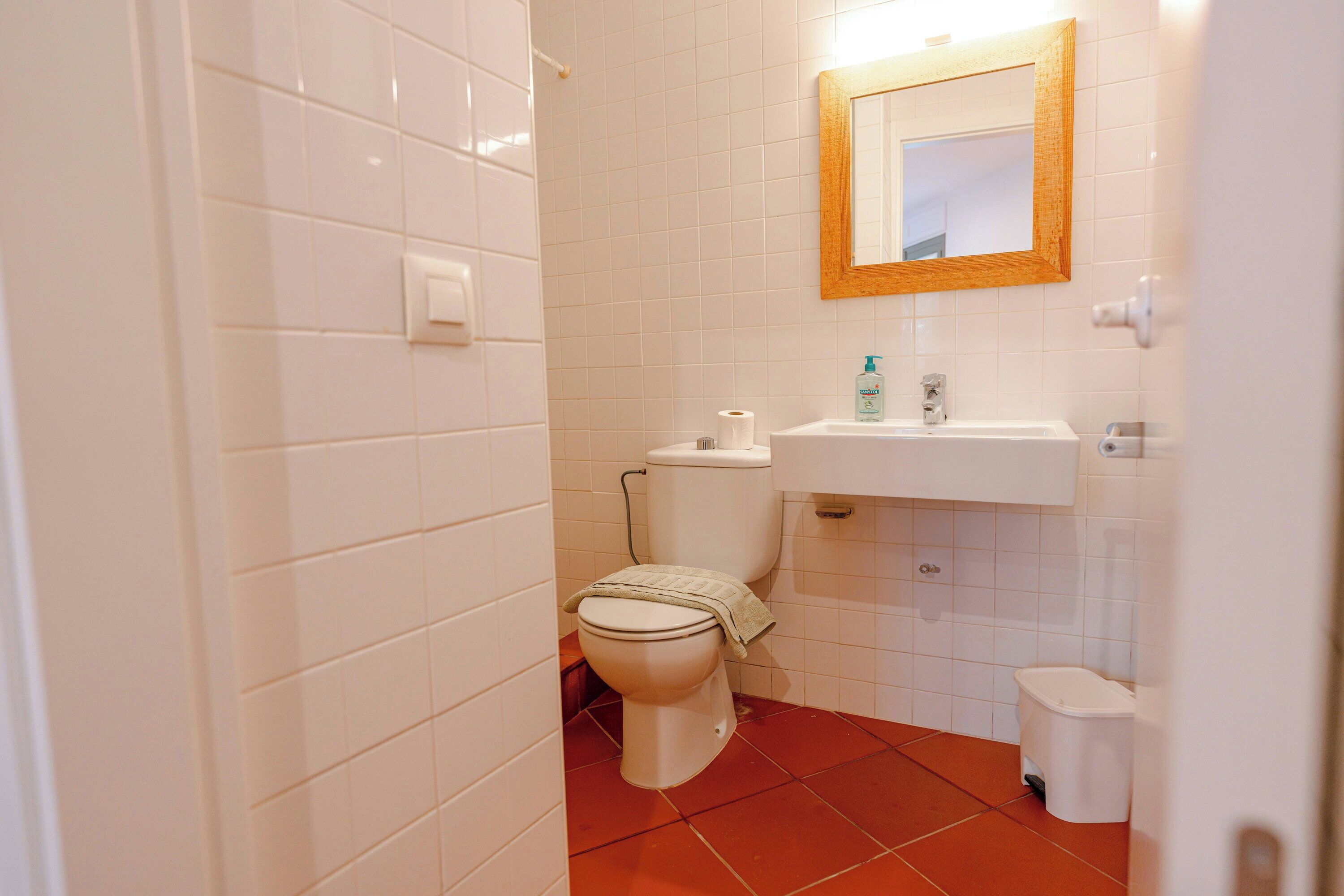 Une salle de bain avec des murs carrelés de carreaux blancs, des toilettes blanches, un lavabo et un miroir avec un cadre en bois.