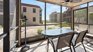 En solbeskinnet terrasse med et glasbord og stole, der vender ud mod en gårdhave med bygninger og grønt.