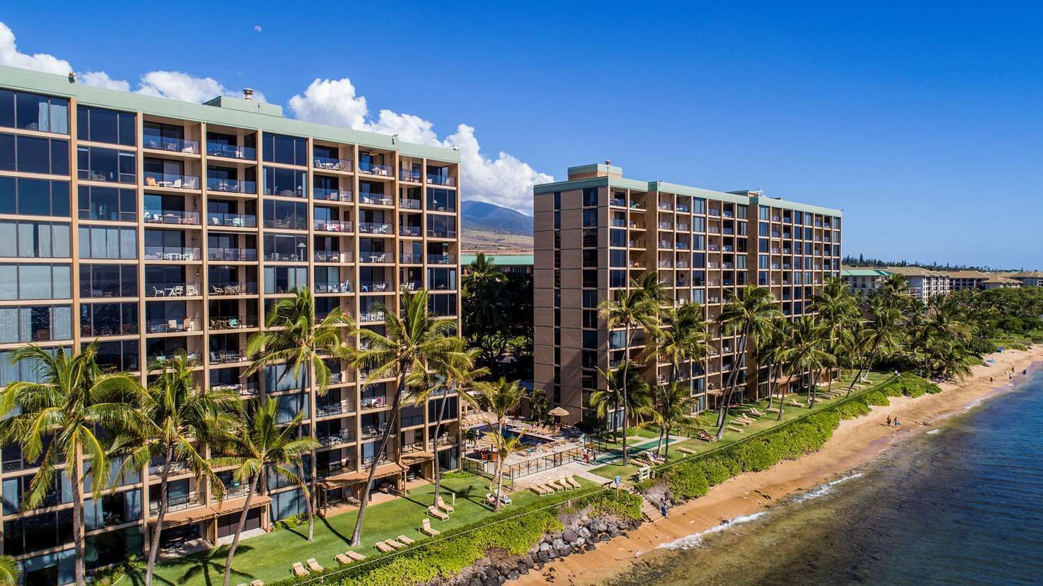 Un hotel con varios pisos, una zona de piscina con sillones de descanso y vista al mar.