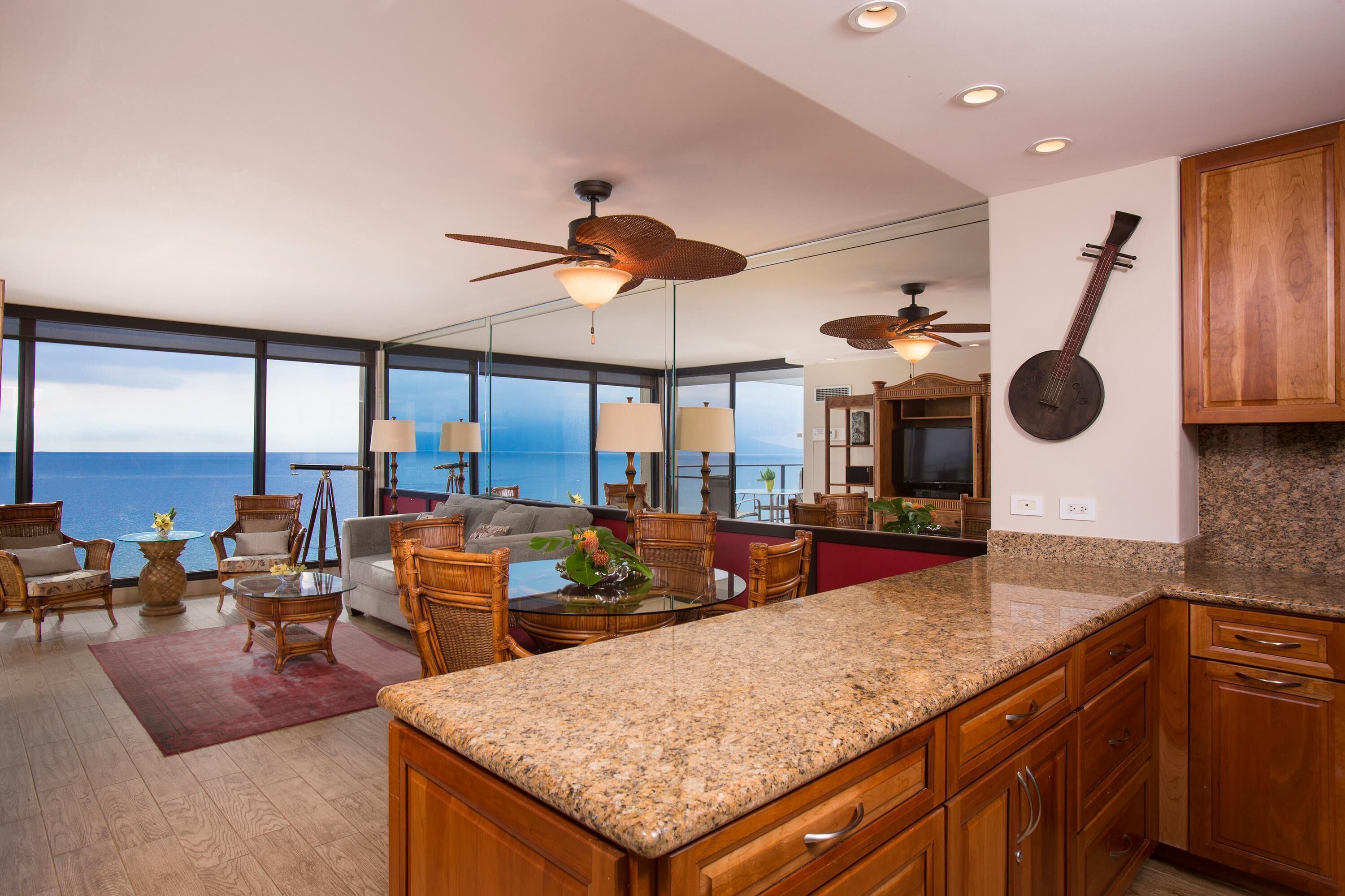 A spacious kitchen with granite countertops, wooden cabinets, and a large island. There is a dining area with wicker furniture and a view of the ocean.