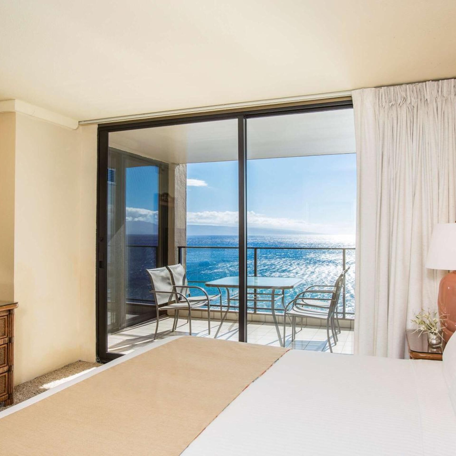 A hotel room with a bed, a glass table with chairs, and a view of the ocean.